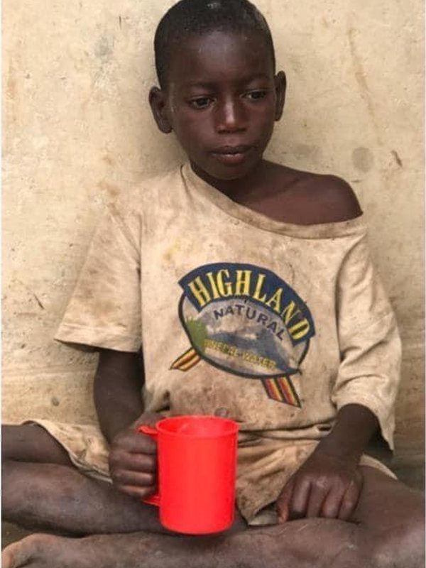 A boy sitting barefoot against a wall in a torn, dirty T-shirt, holding a red cup